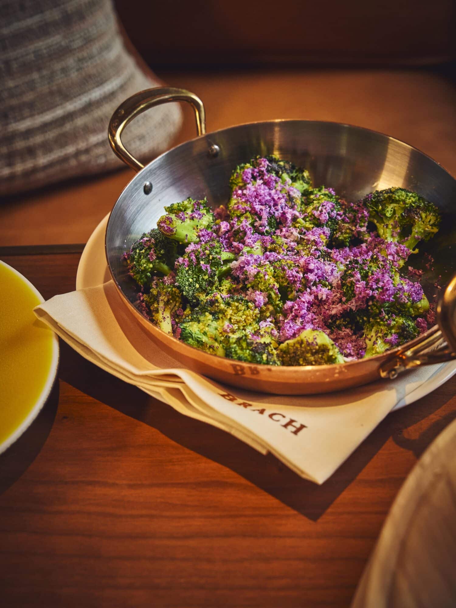 Plat de brocolis rôtis garni de fleurs comestibles violettes, servi dans une poêle en cuivre sur une assiette blanche - GuillaumeCzerw